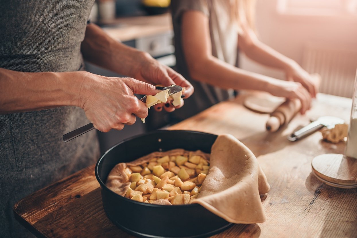 Midsection of person cutting butter into apple pie with another person rolling dough in rustic kitchen